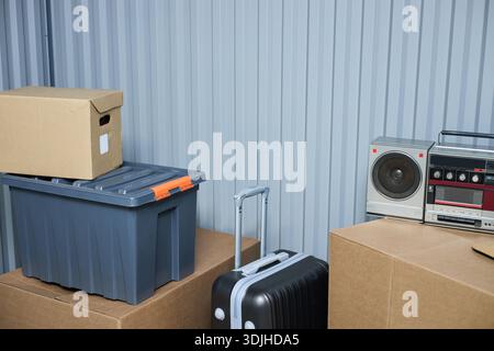 Stacked cardboard boxes, plastic storage container, hard shell suitcase with extended handle, vintage radio and cassette player arranged against corrugated metal wall in storage unit Stock Photo