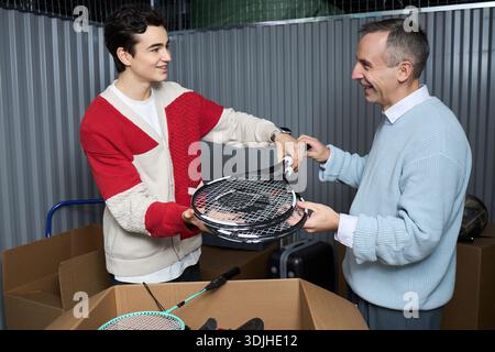 Caucasian teenager boy handing tennis racket to middle aged Caucasian man while unpacking sports equipment from cardboard boxes in storage facility, both smiling and interacting Stock Photo