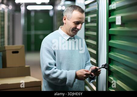 Middle aged Caucasian man unlocking storage unit door using padlock key, standing in hallway lined with green metal doors, cardboard boxes stacked nearby, focused on task Stock Photo