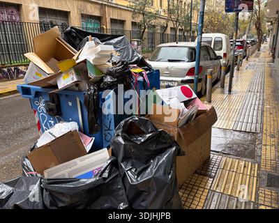 Overflowing blue municipal recycling or refuse bin piled high with cardboard boxes and black rubbish bags on a wet street in Athens, Greece. Urban was Stock Photo