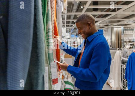 A young African man in a blue jacket examines colorful fabric swatches in a modern retail store. He appears focused and engaged in shopping. Stock Photo