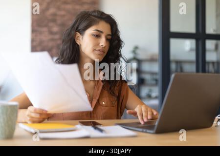 Focused young businesswoman working or studying on laptop, holding documents, sitting at desk indoors, copy space Stock Photo