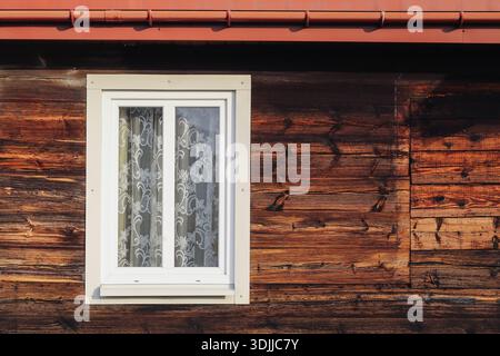 Rustic wooden house wall with white framed window and lace curtain. Traditional architecture detail showing natural wood texture countryside home. Stock Photo
