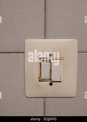 Aged cream colored light switch covered in grime and rust marks, mounted on a vintage tiled wall showing a cracked texture, indicating neglect and a n Stock Photo