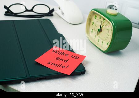 on a white bedside table, a green alarm clock sits next to a tablet and glasses, while a red sticky note reading wake up and smile rests on the device Stock Photo