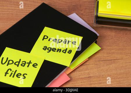 some yellow sticky notes with handwritten reminders, such as prepare agenda, placed on a black notebook on a wooden desk, as part of daily work planni Stock Photo