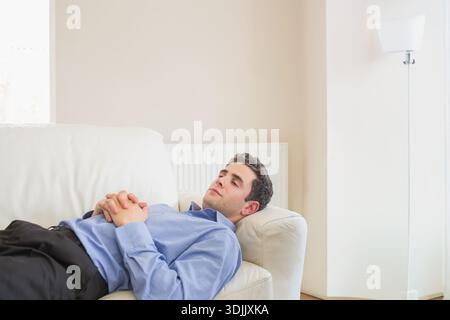 Man wearing light blue button-up shirt reclining on leather sofa in living room, gazing upward. Minimalist, neutral, modern, couch, radiator, lamp, cu Stock Photo