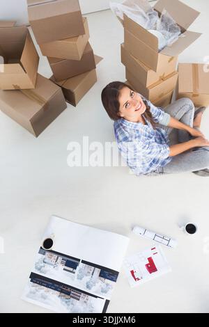 Cardboard moving boxes are surrounding large floor plans on pale floor, showing coffee cups. Unpacking, blueprint, interior, bright, minimalist, home, Stock Photo