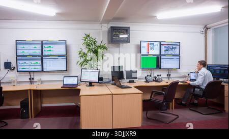 Male operator wearing shirt sitting at desk in control room monitoring screens cables, copy space. Dashboards, displays, monitors, laptops, plant, car Stock Photo