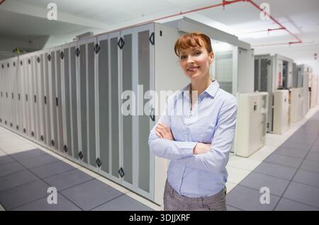 Female technician standing arms crossed in blue shirt gray pants at data center racks, copy space. Serverroom, cabinet, corridor, cooling, electrical, Stock Photo