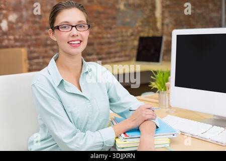 Adult woman working at desk in office wearing green shirt, holding pen on notebooks beside monitor. Workstation, brickwall, plant, glasses, modern, or Stock Photo
