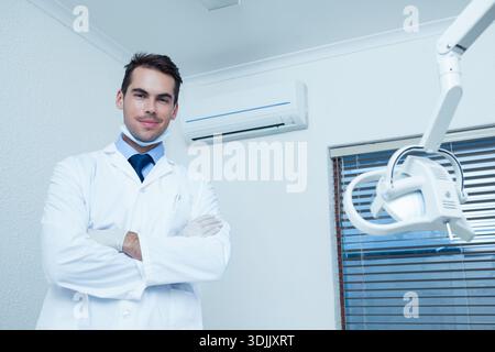 Male dentist standing arms crossed in clinic wearing white lab coat near dental lamp, copy space. Clinical, professional, sterile, medical, overhead, Stock Photo