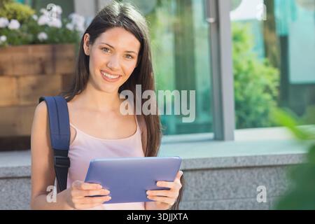 Woman standing beside stone ledge outside modern building, holding purple tablet and backpack strap. Student, campus, device, bag, outdoors, daylight, Stock Photo