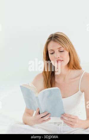 Woman sitting on bed in pale room, wearing camisole and pajamas, holding light blue paperback book. Reading, minimal, serene, softlight, pastel, relax Stock Photo