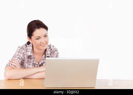 Female wearing plaid shirt leaning on wood table crossing arms looking at silver laptop, copy space. Studio, bright, minimalist, workspace, profession Stock Photo