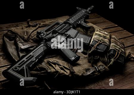 Tactical assault rifle with magazines on a military bag over a wooden surface. Stock Photo
