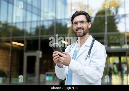 Confident male doctor smiling and using a smartphone for digital healthcare, connecting with patients through telecommunication technology outside a modern clinic building Stock Photo