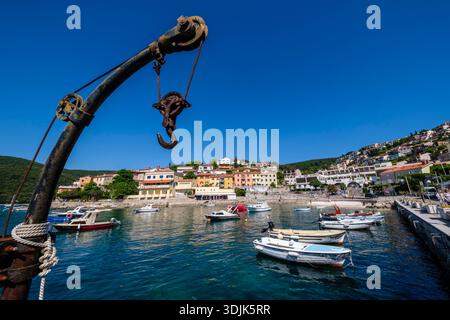 An old rusty crane hook in the foreground with the harbor of Rabac and waterfront buildings. Forested hills are visible in the distance. Stock Photo