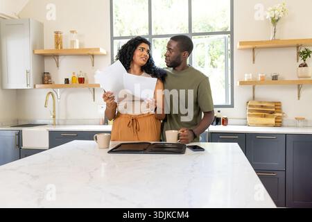 Diverse couple standing behind marble island in casual wear holding papers and mug, checking phone Stock Photo