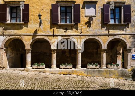 The historic loggia of Grožnjan with its stone arches and pillars. Flowers in planters sit on the base under a yellow facade with dark shutters. Stock Photo