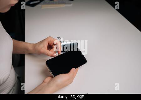 Young woman inserting sim card into new smartphone, with another phone and sim ejector tool nearby on white table, during mobile device setup process Stock Photo