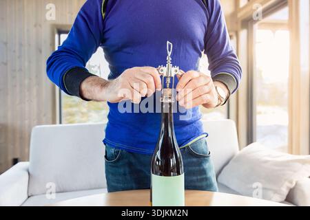 Man in blue sweater is opening wine bottle with corkscrew on wooden table in a bright living room, surrounded by soft furnishings and natural light Stock Photo
