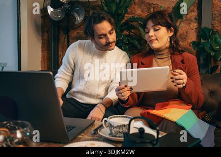 Young couple sitting on a sofa and using a tablet and laptop together in a cozy indoor environment, sharing digital content and interacting in an info Stock Photo