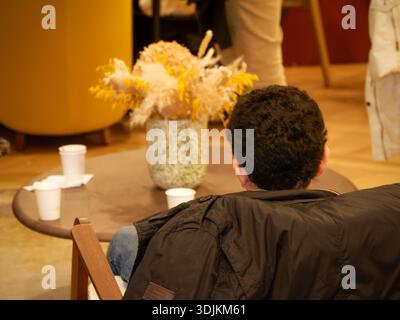 Man sitting in a chair at a café or lounge area, seen from behind, with disposable coffee cups and a floral arrangement on a small table. Stock Photo