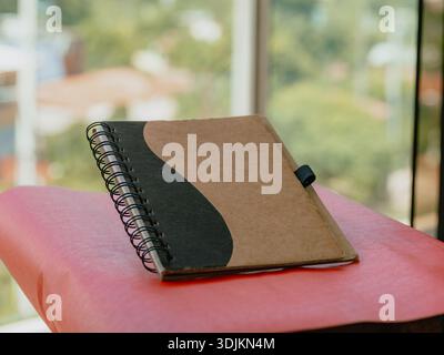 Photograph of a spiral notebook with a brown cover resting on a red surface, with a blurred background and natural light. Useful for education content Stock Photo