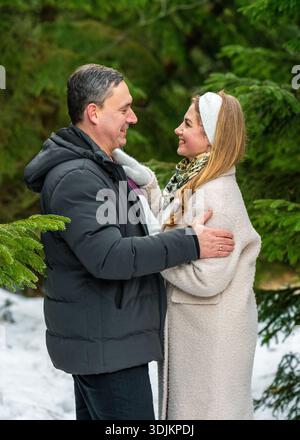 Romantic winter scene of happy couple in snowy coniferous forest. Smiling woman tenderly holds her partner as they gaze into each other eyes. Stock Photo