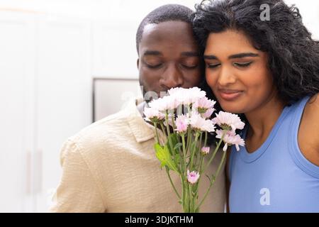 Diverse couple standing holding pink mum bouquet in kitchen wearing beige shirt and blue tank top Stock Photo