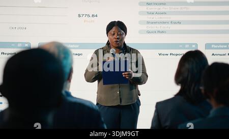 African american woman makes a speech at international summit in conference hall, presentation with reports and investments for networking. Speaker engages audience in business forum. Stock Photo