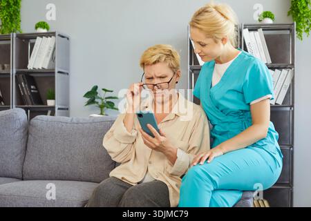 Senior woman using mobile phone with help of caregiver at home, old lady with poor eyesight Stock Photo