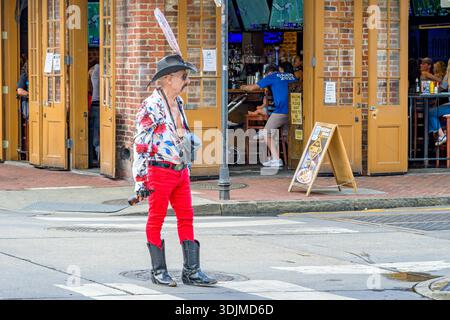 New Orleans, LA, USA - October 18, 2025: Selective focus of colorfully dressed mustachioed man with cowboy hat and boots holding a beer and standing on Bourbon Street, in front of a busy bar, with two polaroid cameras, which he uses to photograph tourists who agree to pose for him. Stock Photo