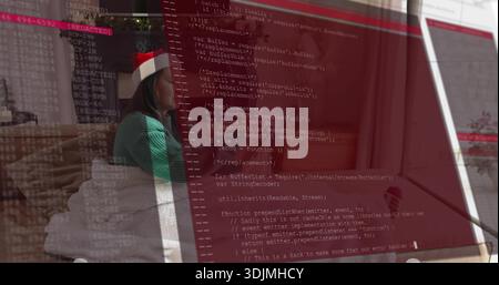 Sitting mature woman with Santa hat and green sweater coding on laptop at home, code overlay Stock Photo