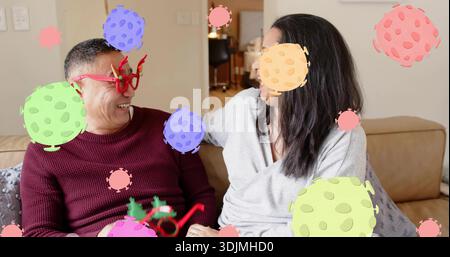 Smiling couple leaning on sofa man wearing burgundy knit red reindeer glasses amid virus overlays Stock Photo