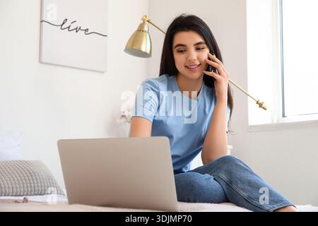 Female freelancer with laptop talking by mobile phone in bedroom Stock Photo