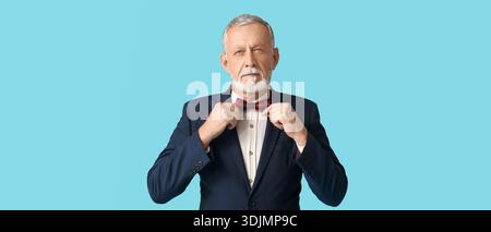 Handsome mature man in stylish formal suit adjusting bow tie on blue background Stock Photo