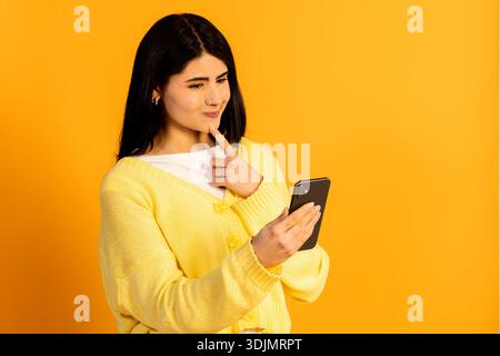 Young woman contemplating a choice or decision, holding a smartphone and resting her finger on her chin with a thoughtful expression on a yellow backg Stock Photo