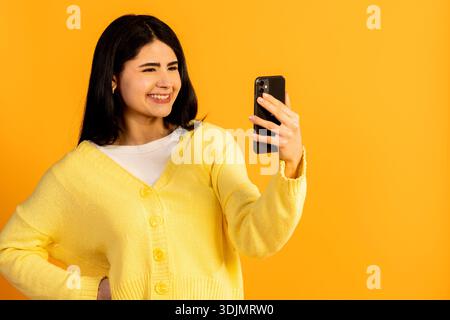 Young woman wearing a yellow cardigan standing in front of a vibrant yellow background, happily interacting with her mobile phone while capturing a pe Stock Photo