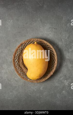 ripe yellow mango in wicker basket on grey textured stone surface, smooth skin fruit in top view with copy space, healthy eating and organic food Stock Photo