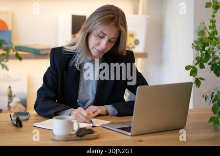 Focused businesswoman working at restaurant, using laptop writing ideas and plans in notebook. Stock Photo