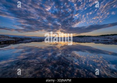 Dramatic sunset over the rocky coast of Pula, with vibrant clouds reflecting in a calm tide pool and a dark treeline in the distance. Stock Photo