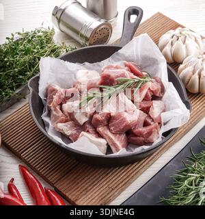 A close-up overhead shot of diced raw pork meat in a cast iron skillet, seasoned with fresh rosemary. Accompanying ingredients like garlic bulbs, chil Stock Photo