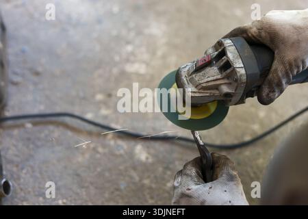 Focused worker in workshop grinds piece of metal with an angle grinder. Dirty gloved hands hold powerful tool and object, process creating sparks on c Stock Photo