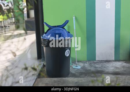 Black trash can with blue lid on concrete floor. This outdoor setting shows mundane scene with green striped wall and leaning mop for cleaning public Stock Photo