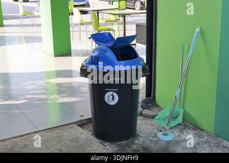 Black plastic trash can with blue lid for garbage disposal and waste management. green dustpan and broom await cleaning, promoting sense of tidiness a Stock Photo