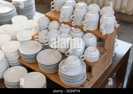 Large group of clean white ceramic dishware, including plates, cups, and bowls, neatly arranged wooden stand. This catering setup for restaurant buffe Stock Photo