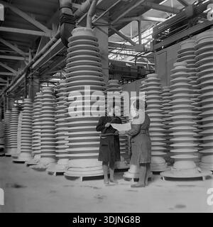 Slavyansk, Donetsk region, Ukraine, USSR. January 1978. Archive photo inside the Slavyansk High-Voltage Insulator Plant (ZVI). Two female workers, likely quality control engineers, stand among rows of massive white porcelain insulators. One woman holds large metal calipers while the other examines a technical blueprint. They are discussing precise manufacturing specifications. The image highlights the scale of Soviet heavy industry and women's role in production during the peaceful era of the Ukrainian SSR. Stock Photo