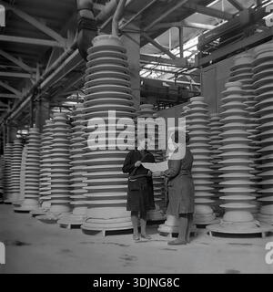 Slavyansk, Donetsk region, Ukraine, USSR. January 1978. Archive photo inside the Slavyansk High-Voltage Insulator Plant (ZVI). Two female workers, likely quality control engineers, stand among rows of massive white porcelain insulators. One woman holds large metal calipers while the other examines a technical blueprint. They are discussing precise manufacturing specifications. The image highlights the scale of Soviet heavy industry and women's role in production during the peaceful era of the Ukrainian SSR. Stock Photo